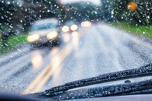 Driver POV (point of view) looking over the windshield wipers through the wet, blurry, partially opaque, partially transparent, spot speckled windshield of a car traveling on a rural highway during an autumn rain storm. Hazy, blurred headlights of approaching vehicle traffic are just blobs of light through the streaking and splattered raindrop water.